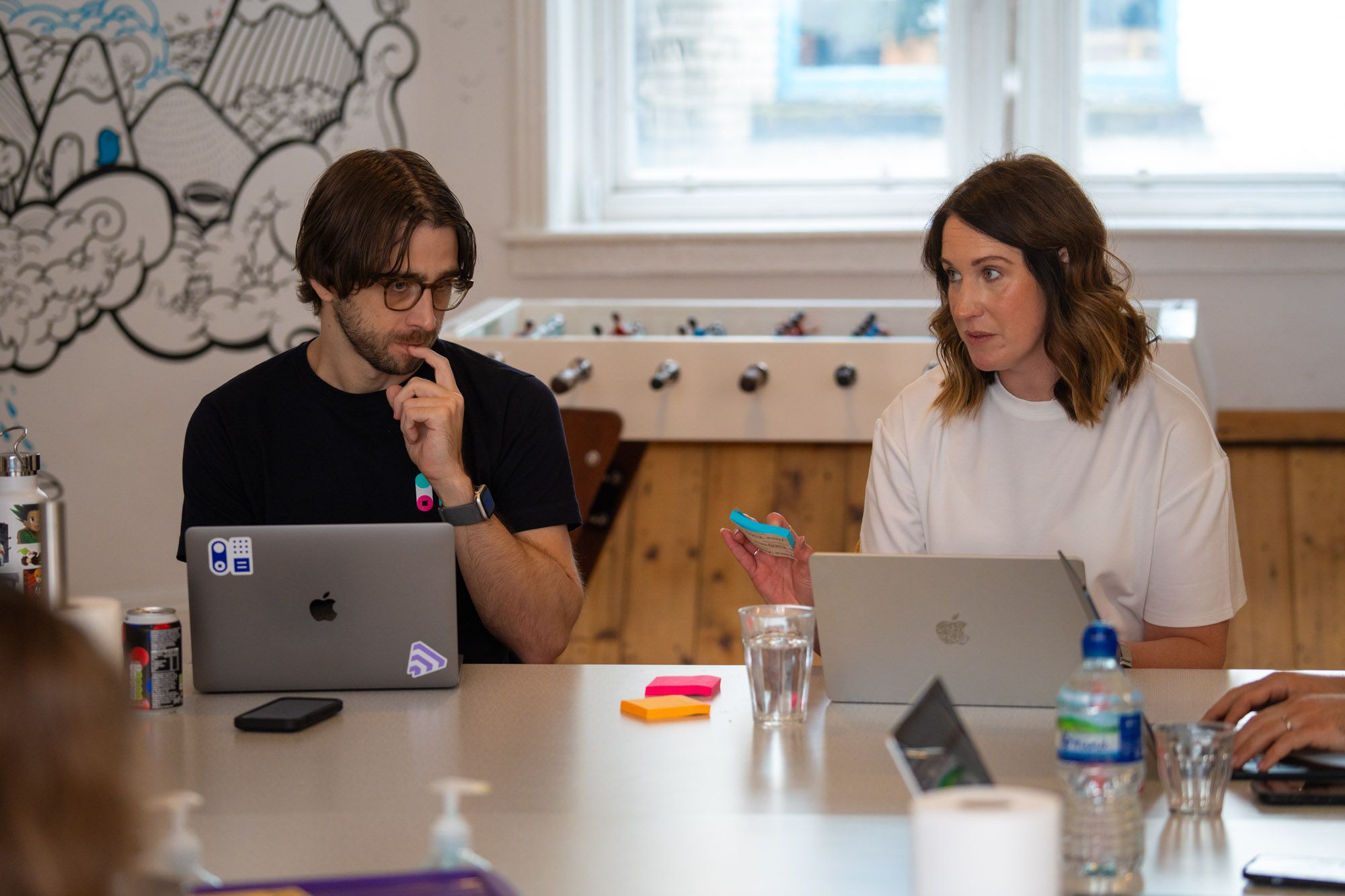 Two people with laptops at a table, a woman speaking while a man listens thoughtfully.