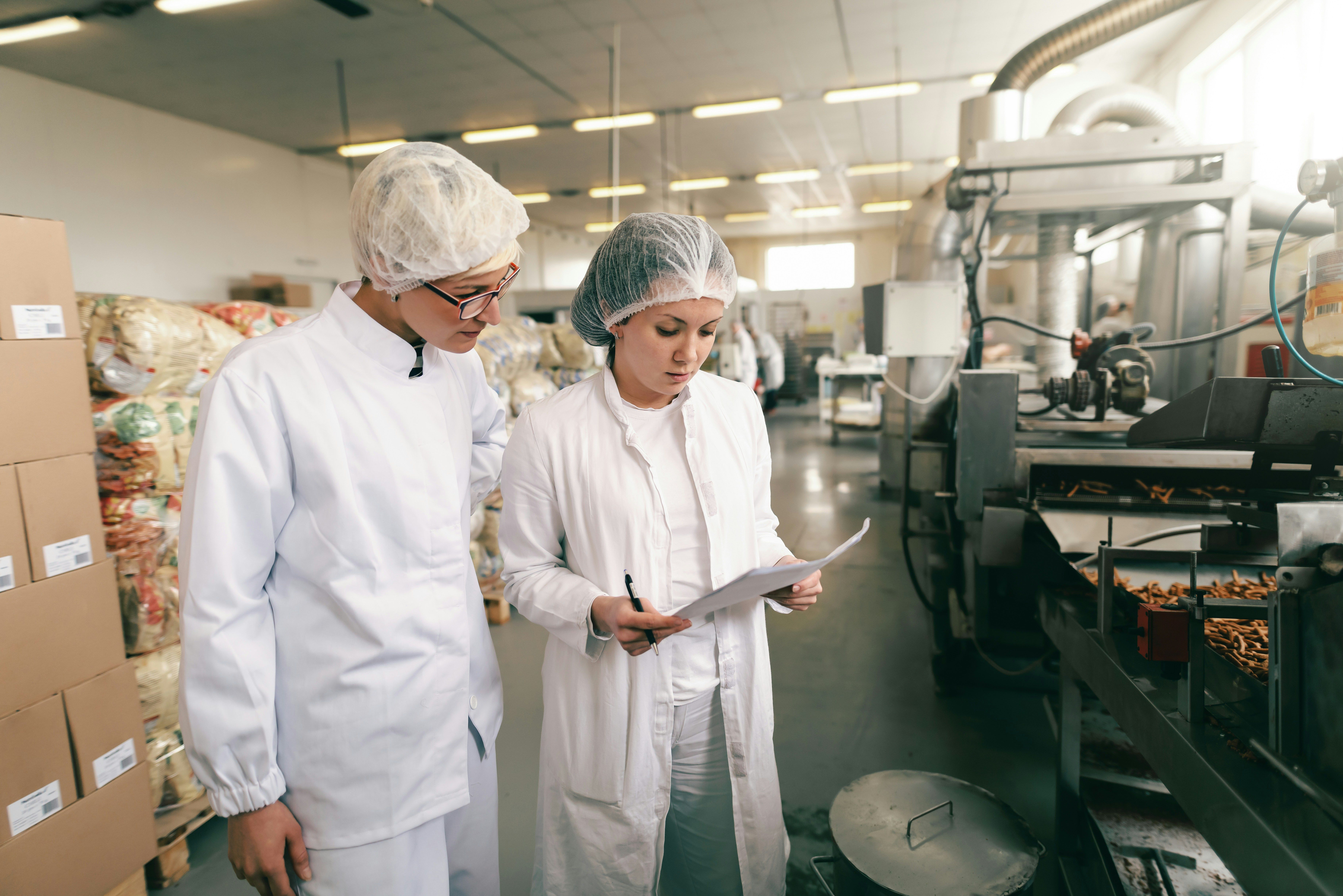 Two women in white protective suits and hairnets examine documents in a food production facility.