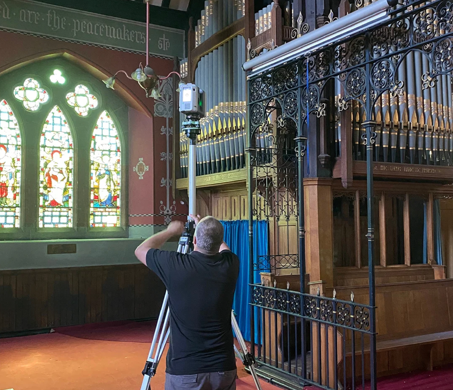 A person adjusts a laser scanner on a tripod inside a church with stained glass windows and a pipe organ.