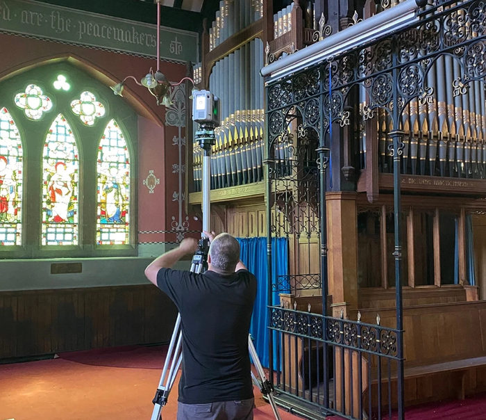 A person adjusts a laser scanner on a tripod inside a church with stained glass windows and a pipe organ.