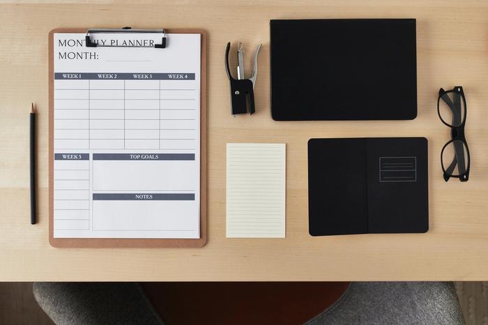 a wooden desk with a clipboard , notebook , glasses , stapler , pencil and a calendar .
