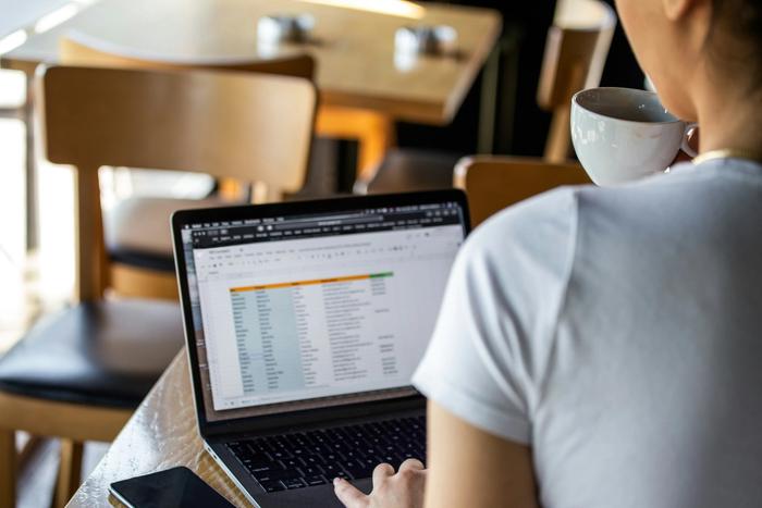 a woman is sitting at a table with a laptop looking at a spreadsheet while holding a cup of coffee