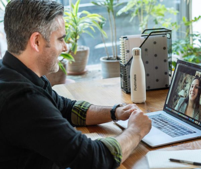 a man sits at a desk with a laptop and a water bottle that says postmates