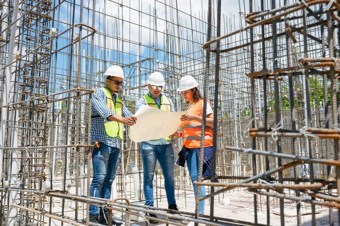 Three construction workers review blueprints on a rebar-filled construction site.