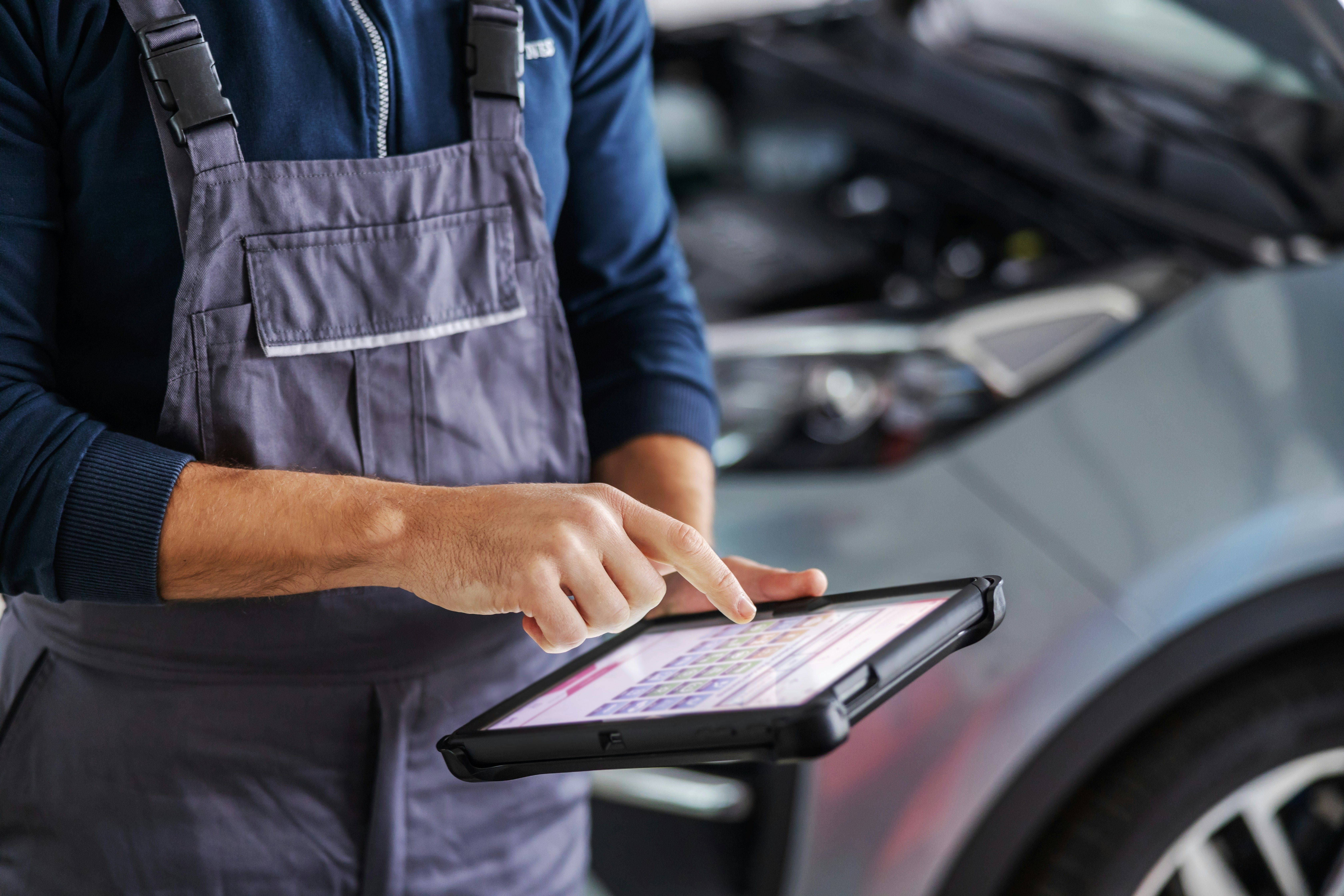 Mechanic in overalls uses a tablet to diagnose a car with its hood open.