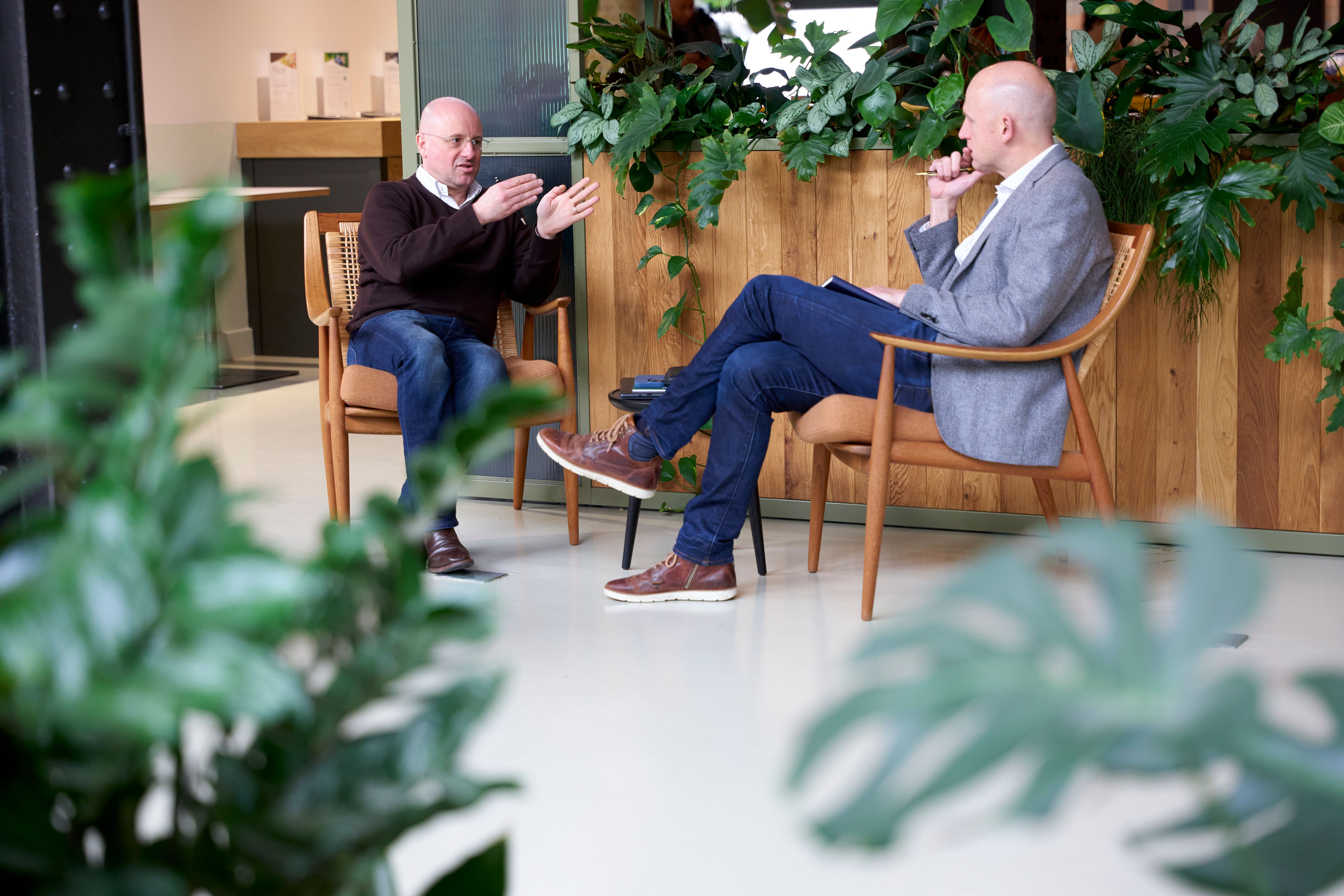 Two men conversing in a modern, plant-filled room, one gesturing, the other taking notes.