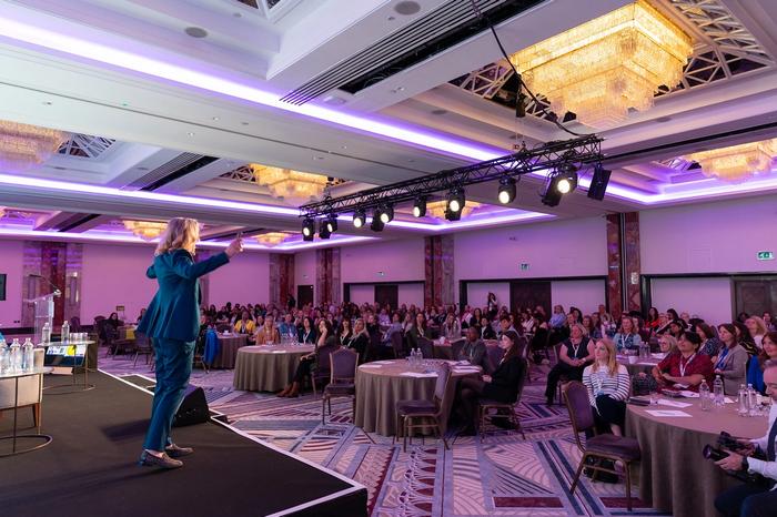Lynda addresses a large audience seated at round tables in an elegant conference room.