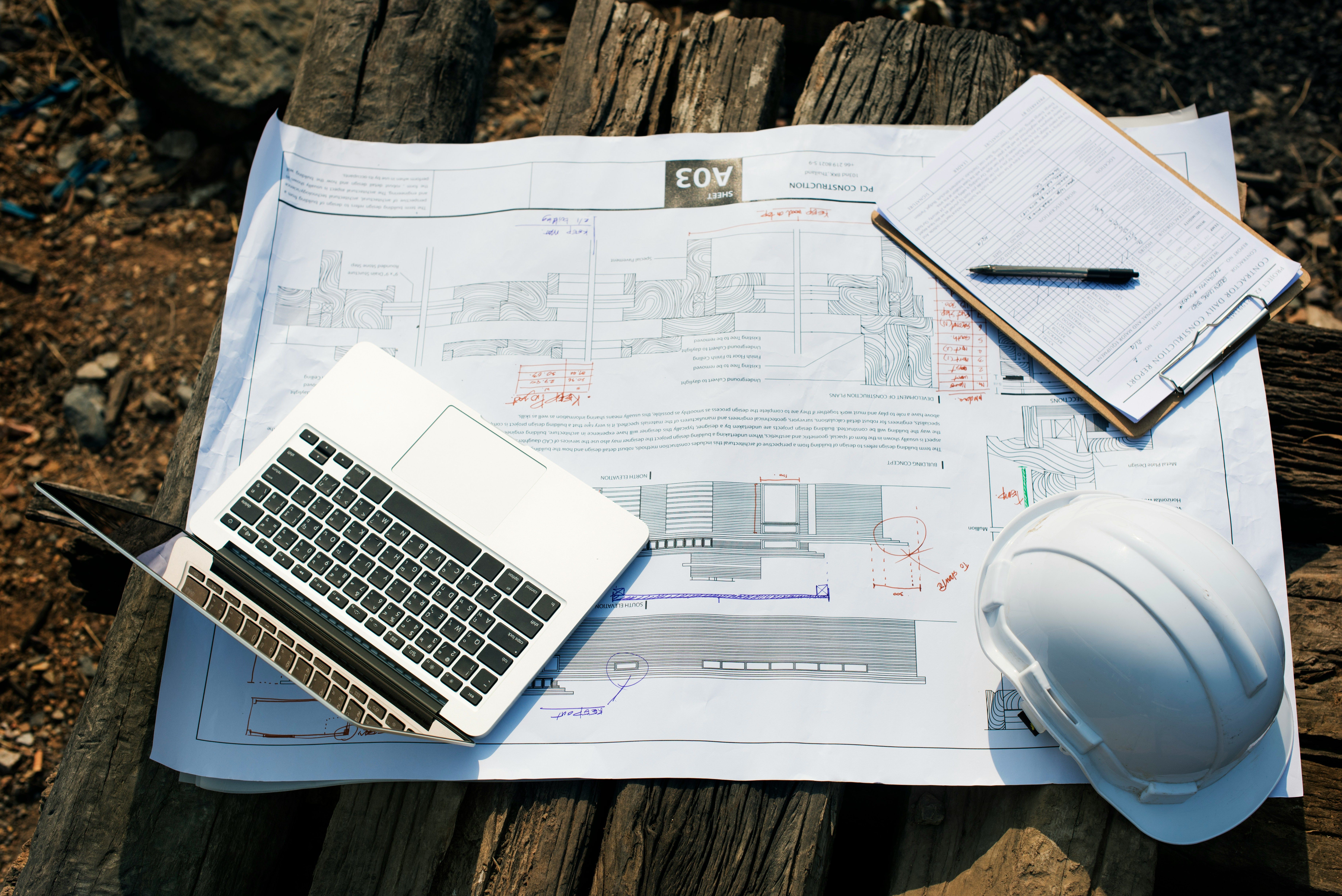 Laptop, hard hat, and clipboard on blueprints atop a wooden surface.