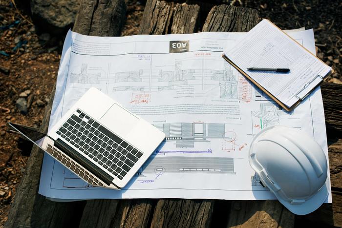Laptop, hard hat, and clipboard on blueprints atop a wooden surface.