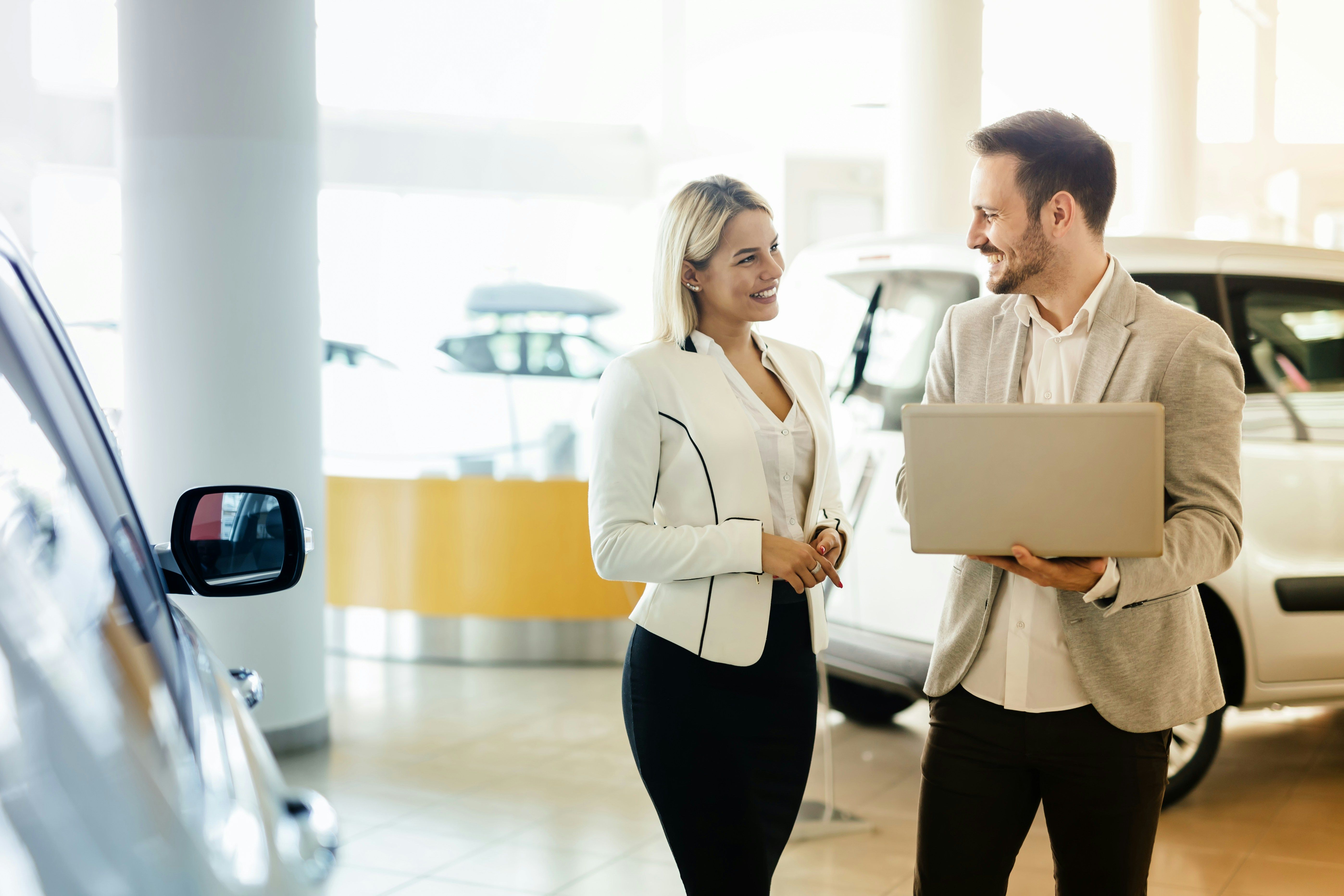 A smiling man holding a laptop talks with a woman in a car showroom.