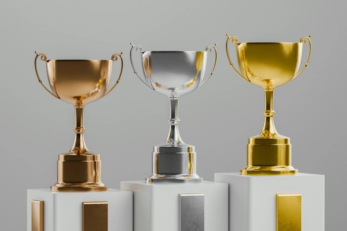 três troféus, de ouro, prata e bronze, estão sobre blocos brancos