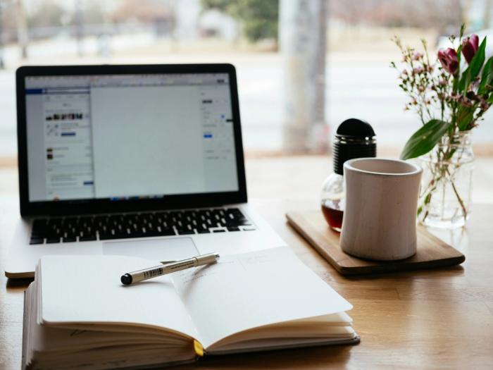 a laptop computer , a notebook , a pen , and a cup of coffee are on a wooden table .