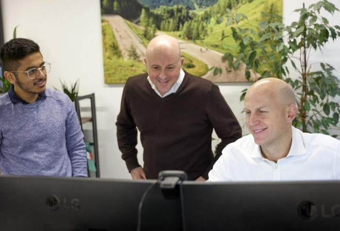 Three men smiling while looking at computer screens in an office.