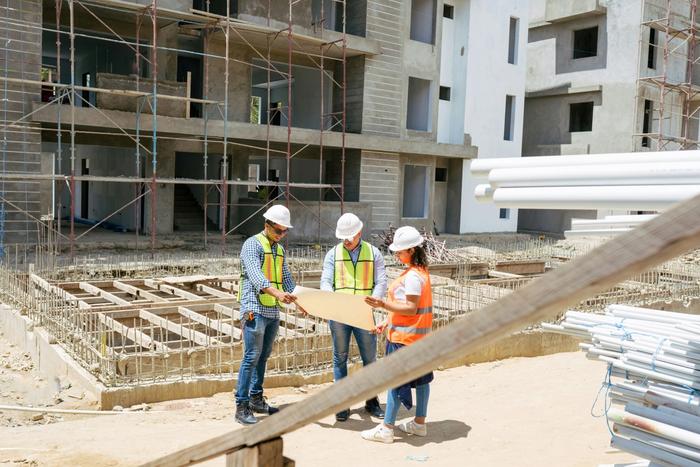 Three construction workers in hard hats review plans on a building site.