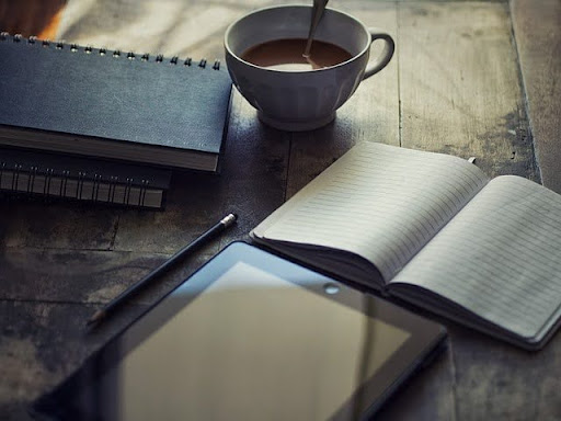a cup of coffee sits next to an open notebook and a tablet