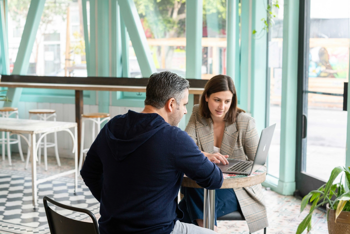 a man and a woman sit at a table looking at a laptop