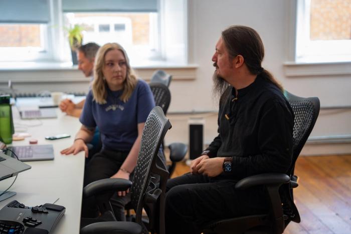 A man with long hair and beard talks to a woman at a desk in an office.