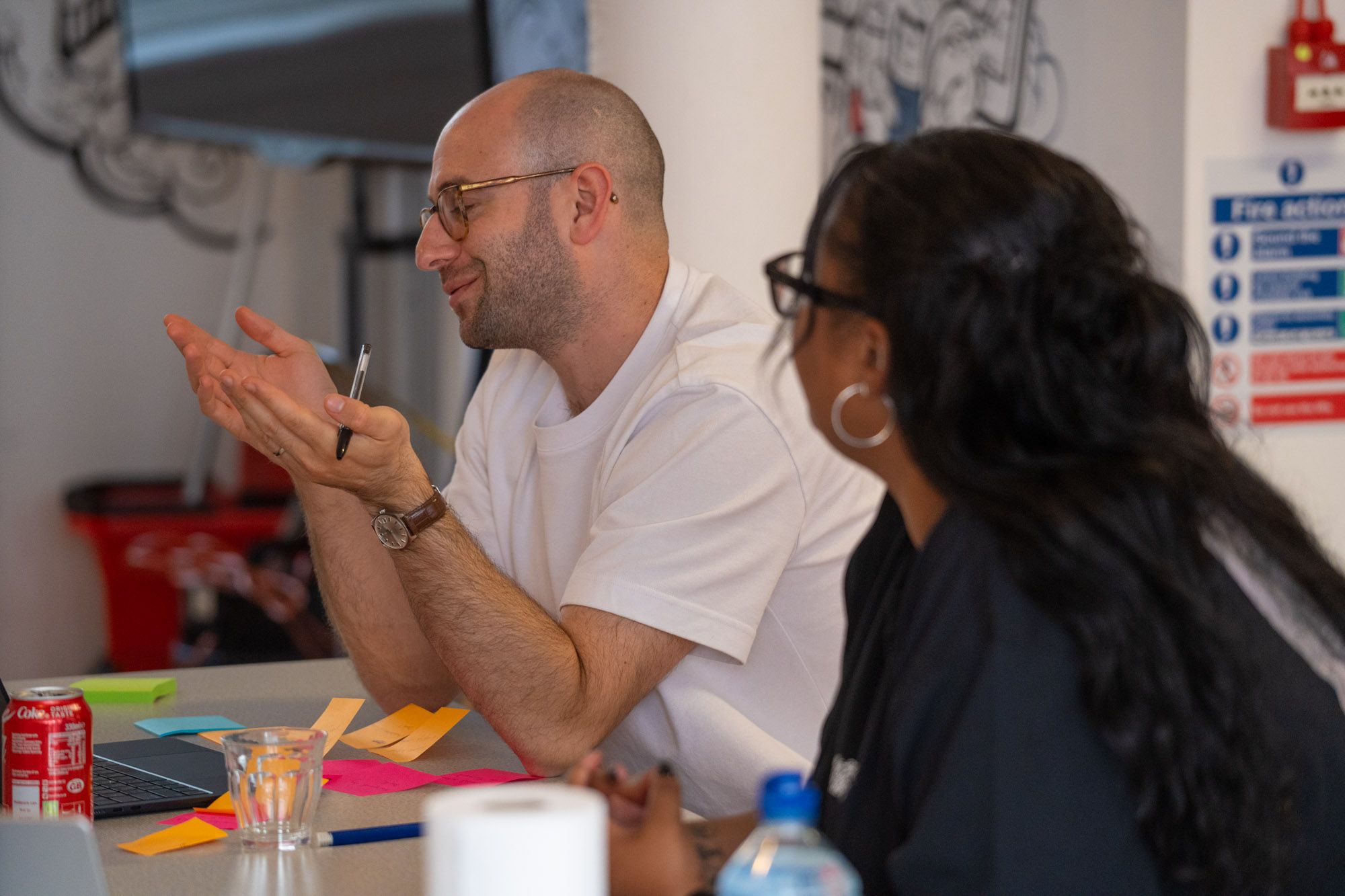 A man in glasses gestures with his hands while speaking to a woman at a table with a laptop and sticky notes.