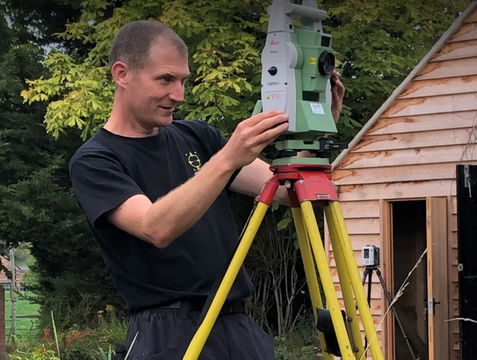 A man operates a surveying total station on a tripod outdoors.