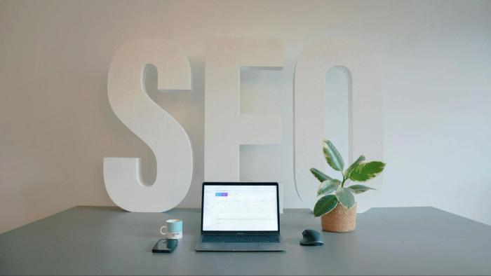 Minimalist desk setup with a laptop showing analytics, a coffee mug, and a plant, with large freestanding letters spelling “SEO” in the background.