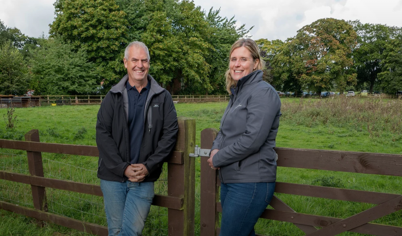 A smiling man and woman stand by a wooden fence in a field.