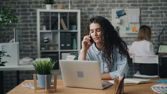 A woman sits at a desk looking stressed while she reviews spreadsheets and makes business phone calls.