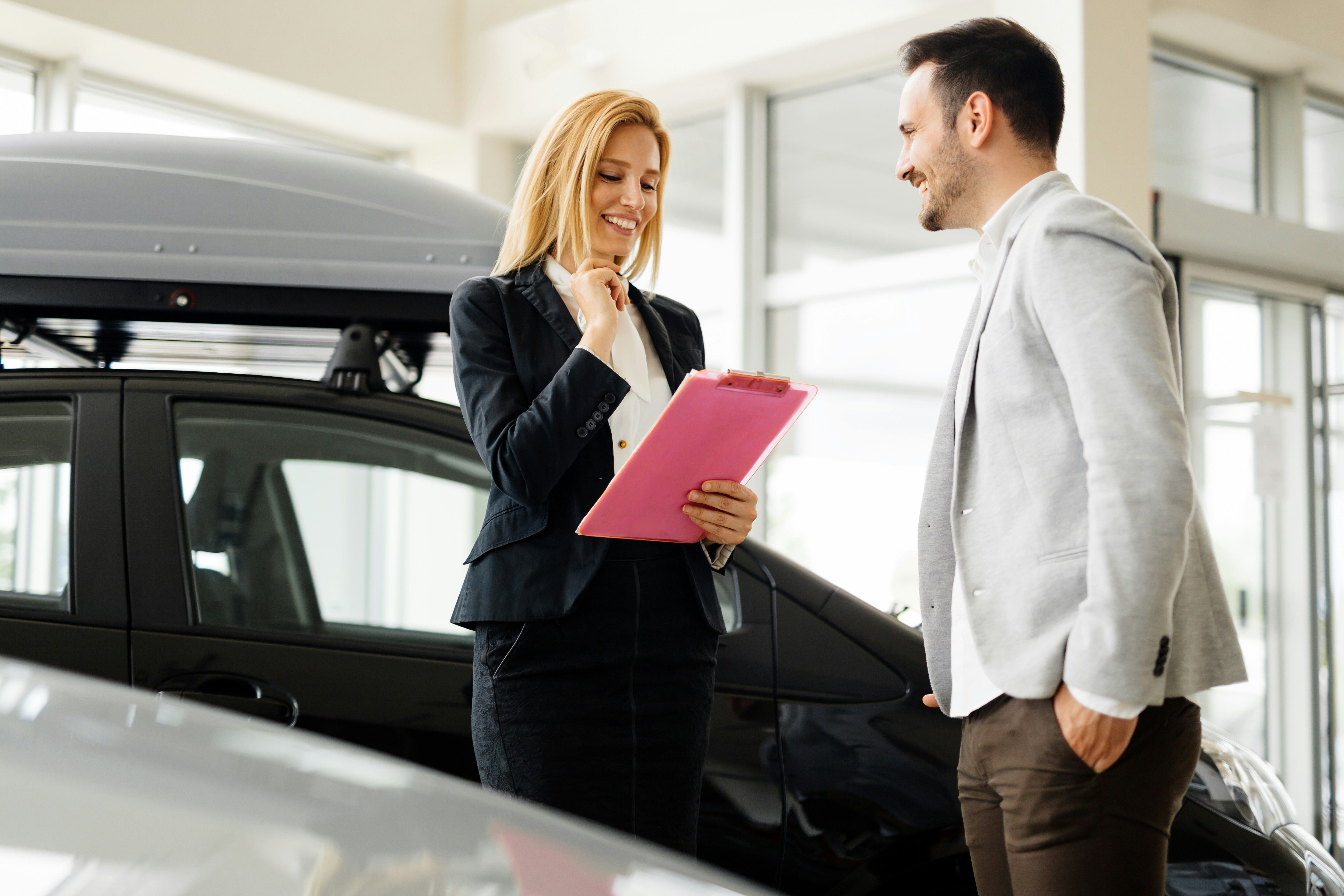 A smiling car salesperson holding a clipboard talks with a male customer in a car showroom.