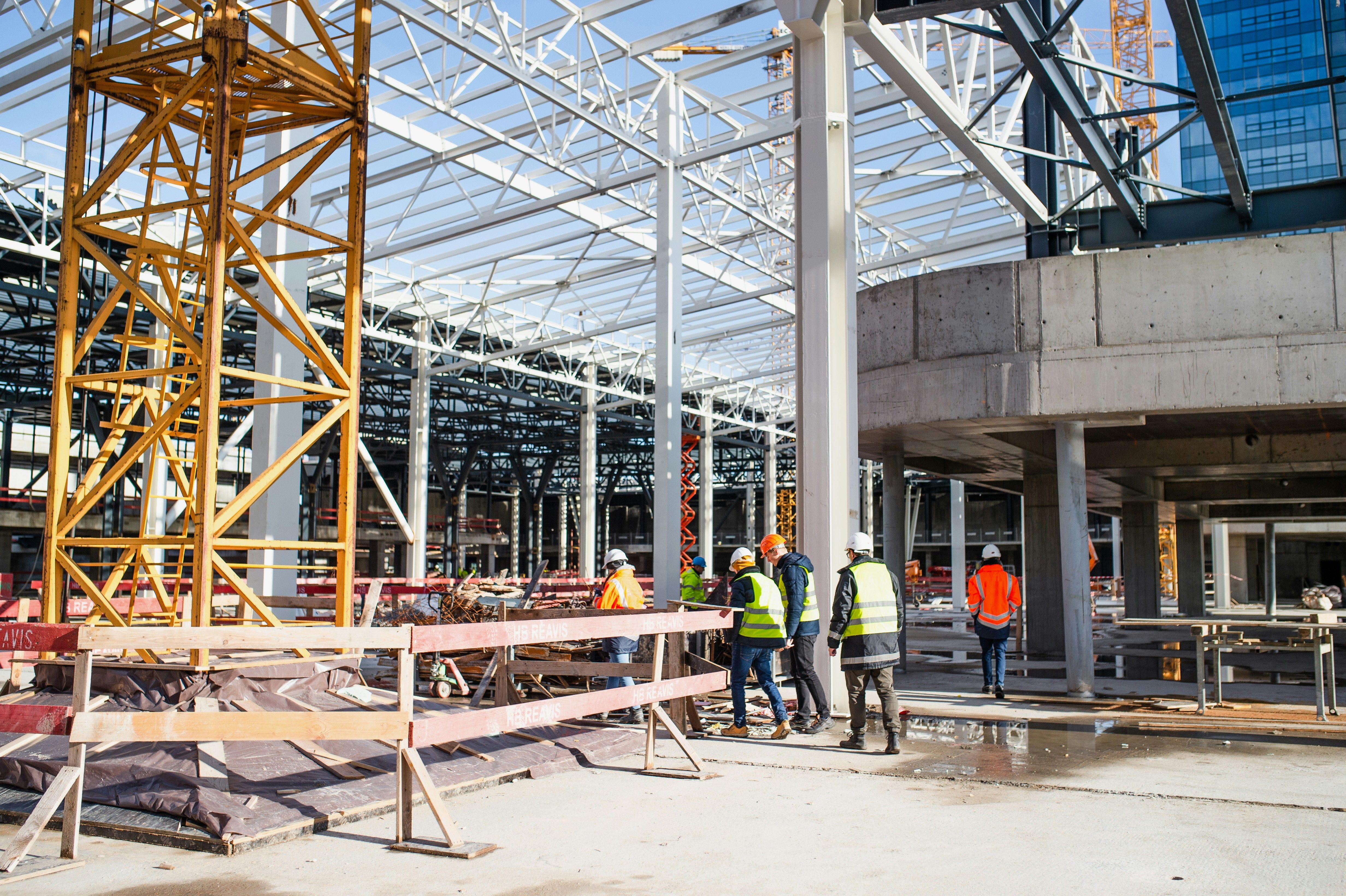 Um grande edifício em construção, com estrutura de aço, elementos de concreto, um guindaste amarelo e vários trabalhadores usando equipamentos de segurança.