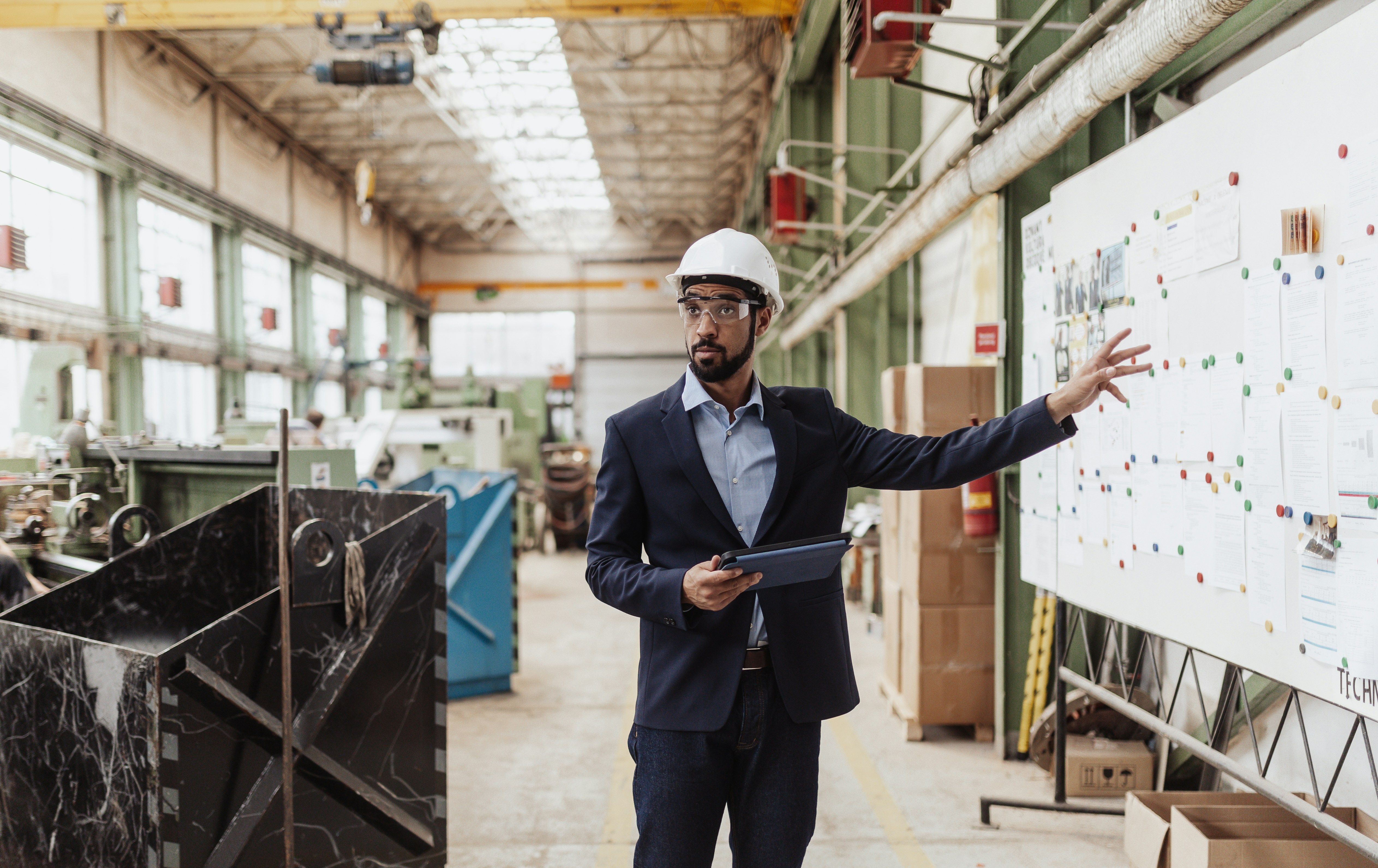 An engineer in a hard hat and safety glasses holds a tablet while pointing at a planning board in a factory.
