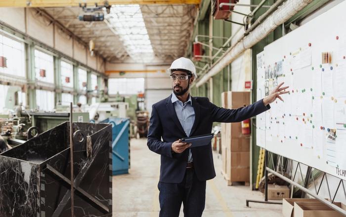 An engineer in a hard hat and safety glasses holds a tablet while pointing at a planning board in a factory.