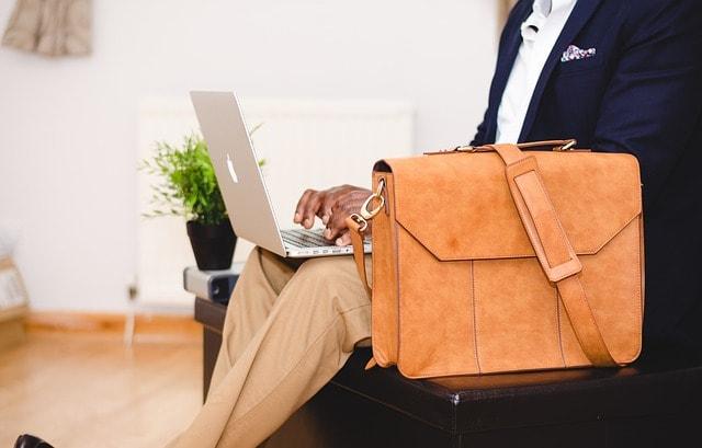 A man is sitting on a couch using a laptop and a briefcase.