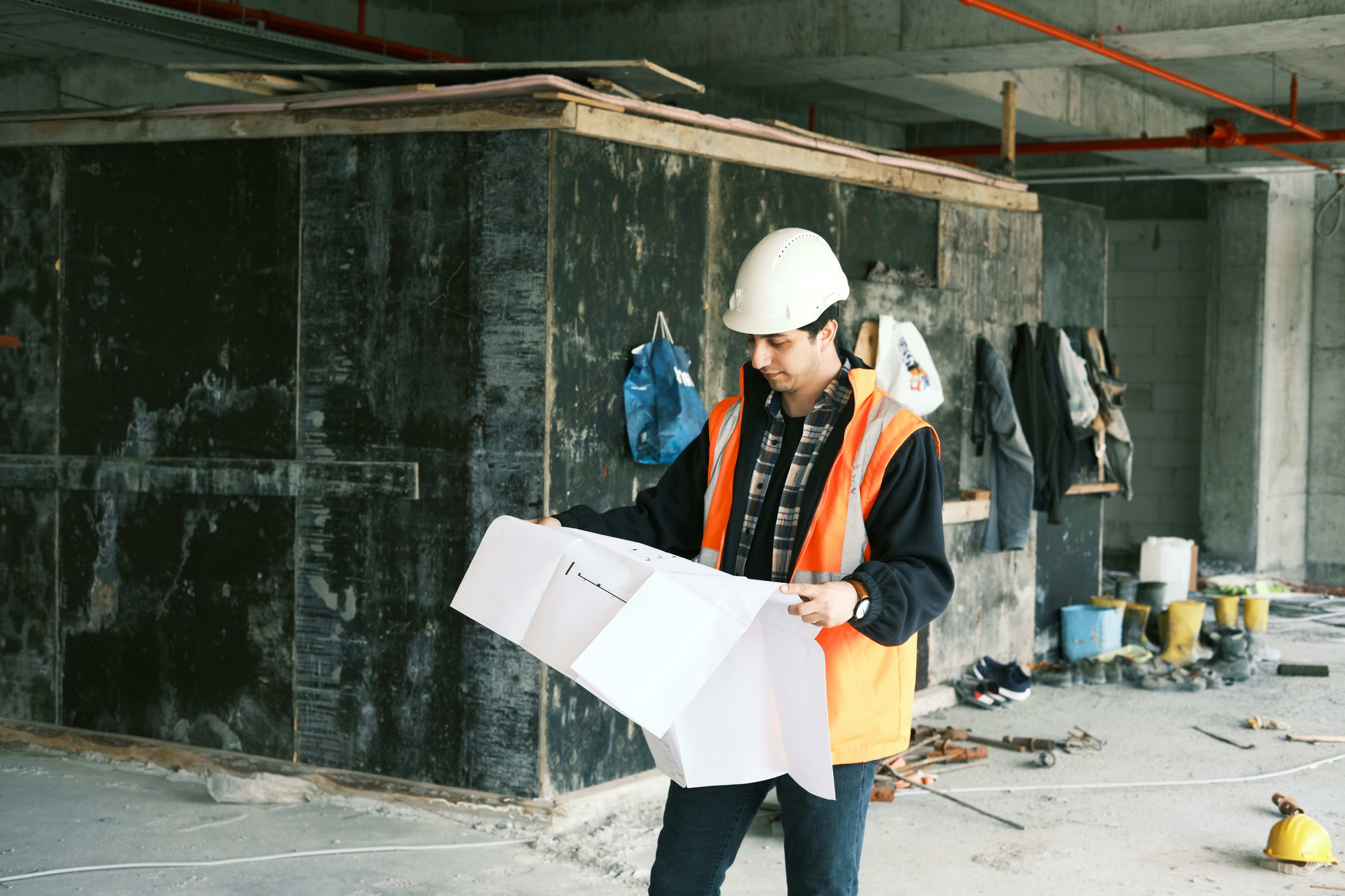 A construction worker in a hard hat and safety vest reviews blueprints on a job site.