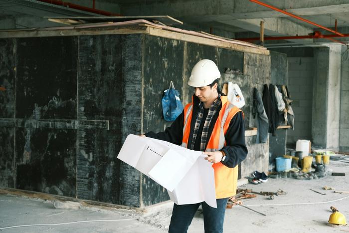 A construction worker in a hard hat and safety vest reviews blueprints on a job site.