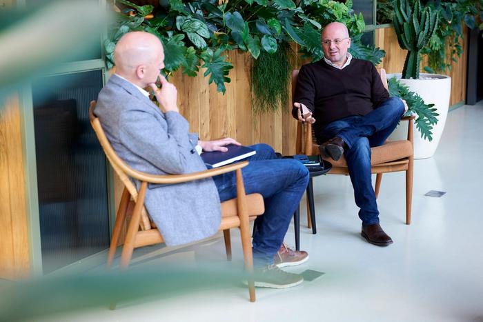 Two men in conversation, one listening and one talking, in a modern office with indoor plants.