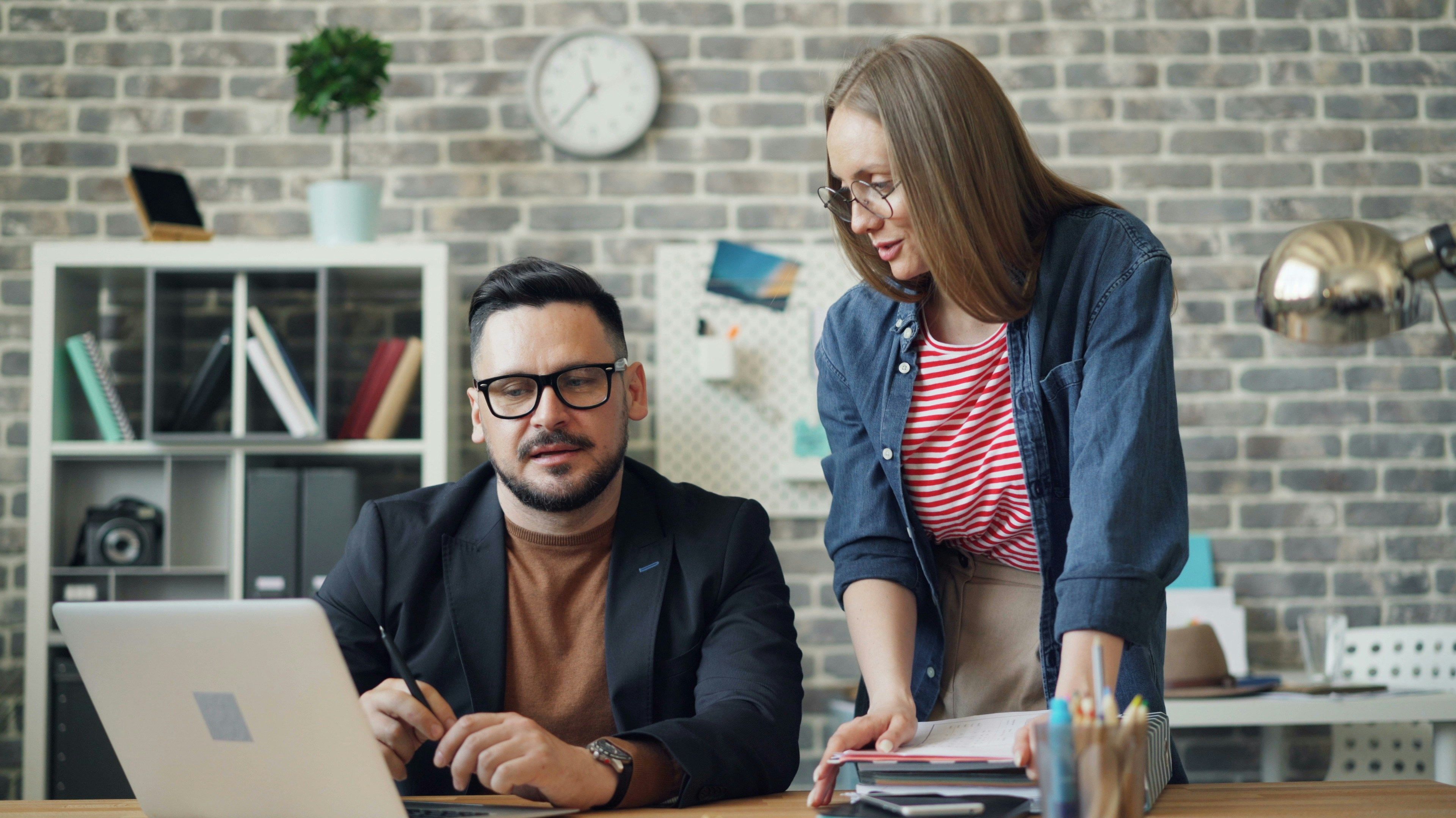 A man sat at a desk while a woman stands over him looking at his laptop