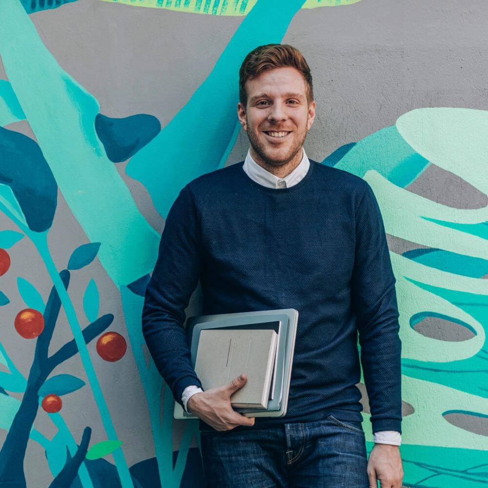 Man standing in front of a colourful wall with a laptop and notebook under his arm