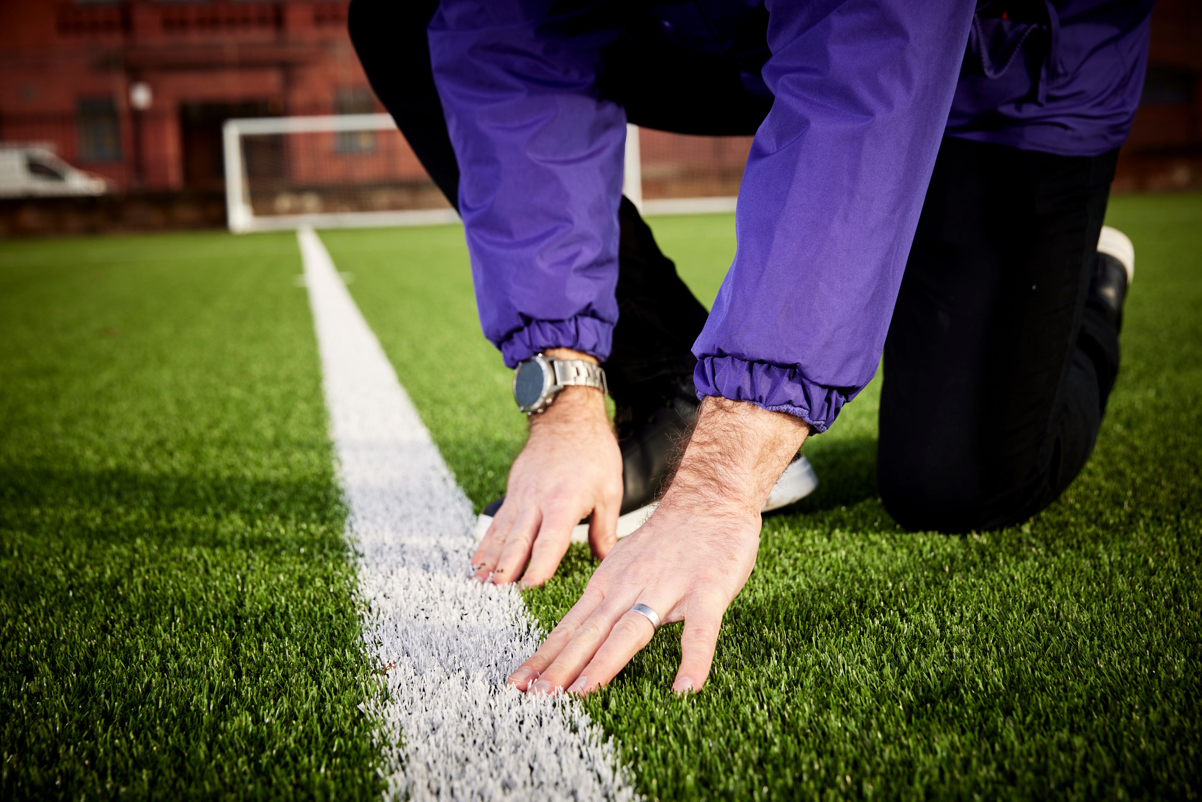 A person in a purple jacket kneels on a green sports field, hands touching a white line.