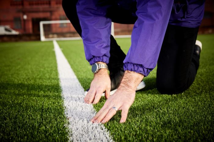 A person in a purple jacket kneels on a green sports field, hands touching a white line.