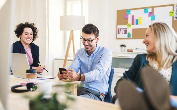 a group of people are sitting at a table in an office looking at a cell phone .