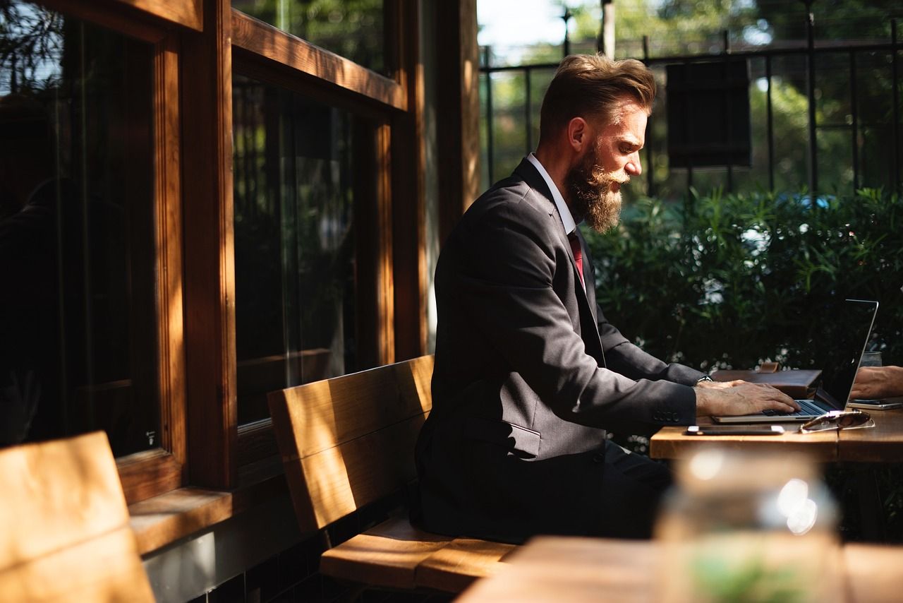 A bearded man in a suit works on a laptop at an outdoor cafe.