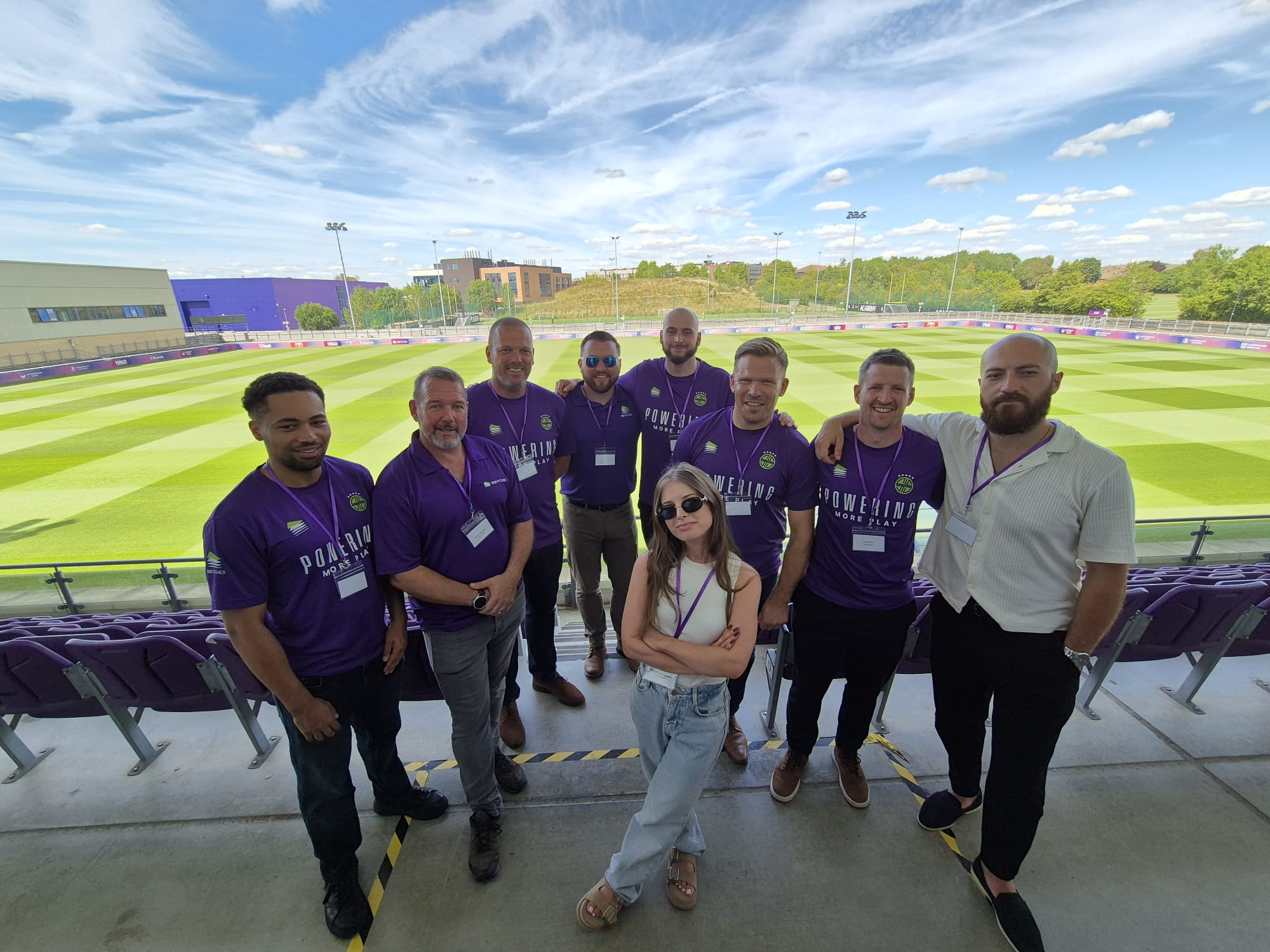 Nine people, mostly men in purple shirts with "POWERING YOUR TEAM" text, pose in stadium seats overlooking a green sports field.