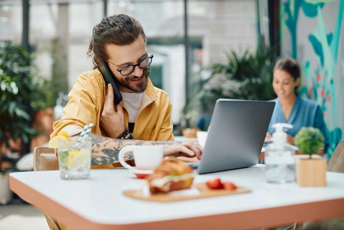 a man is sitting at a table with a laptop and talking on a cell phone .