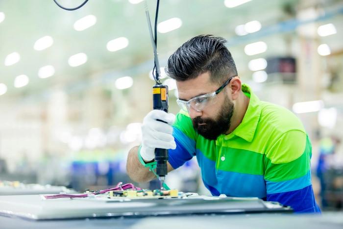 A man wearing safety glasses and gloves uses a power screwdriver to assemble electronics in a factory.