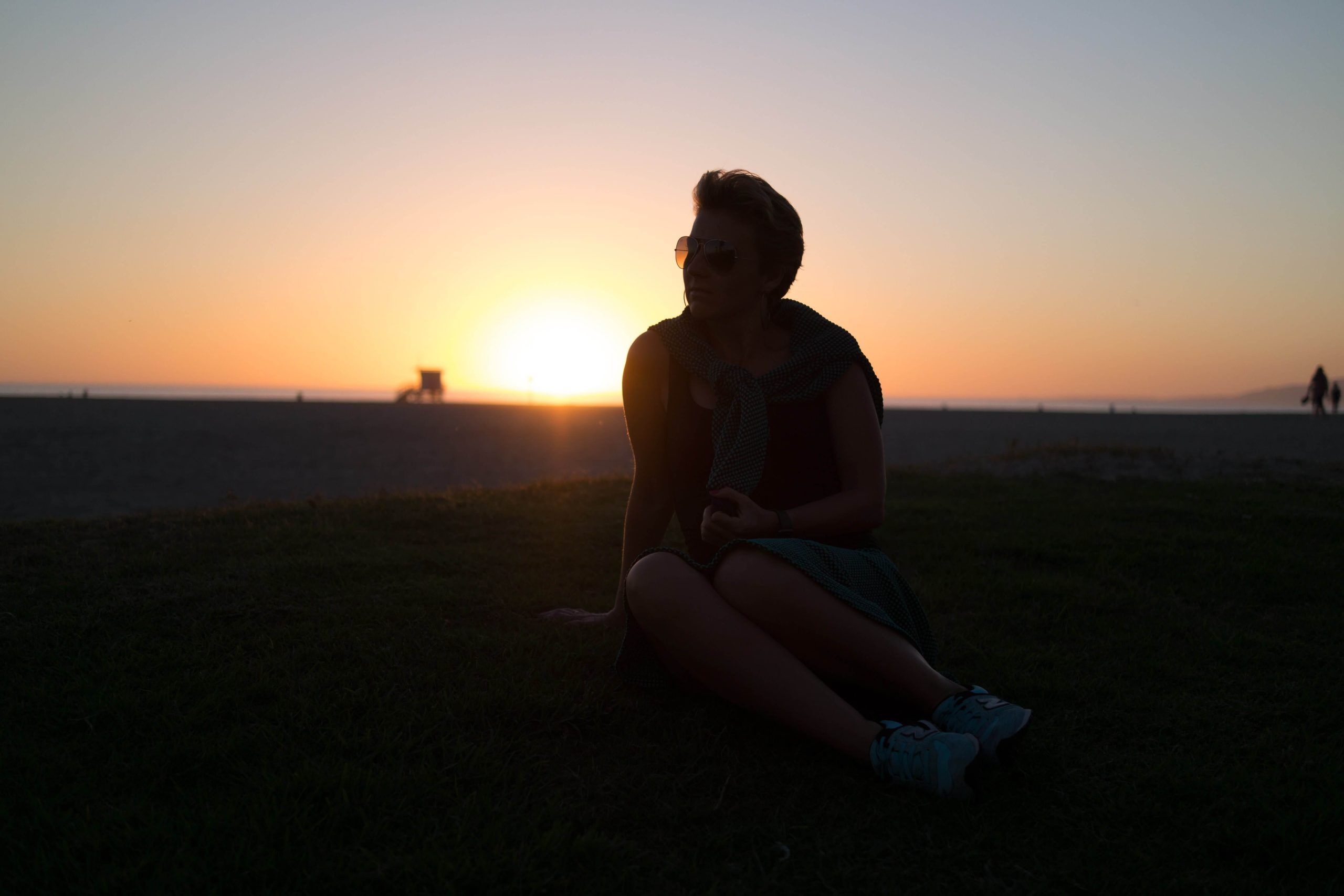 Woman wearing sunglasses sits on grass beside a beach at sunset