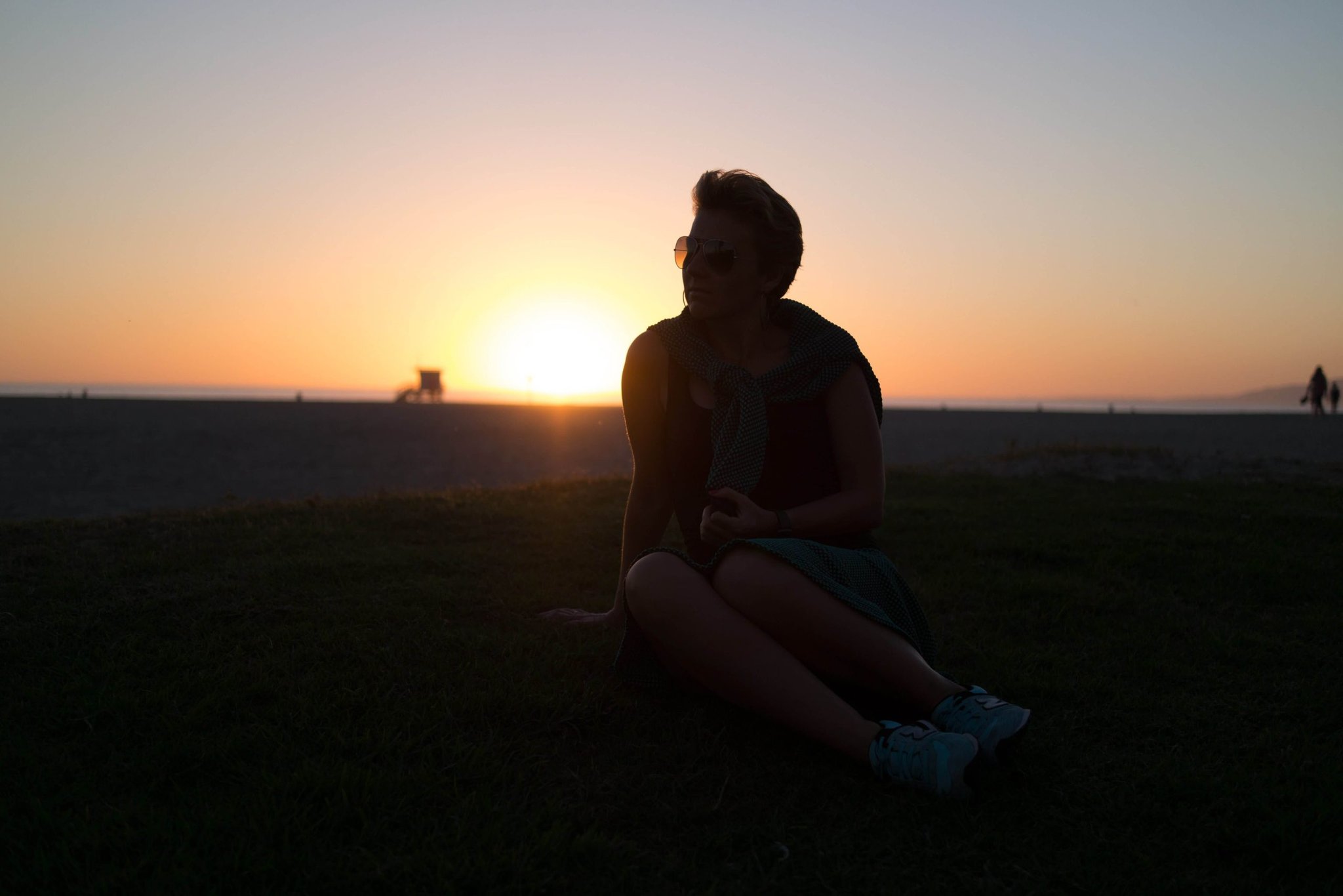 Woman wearing sunglasses sits on grass beside a beach at sunset