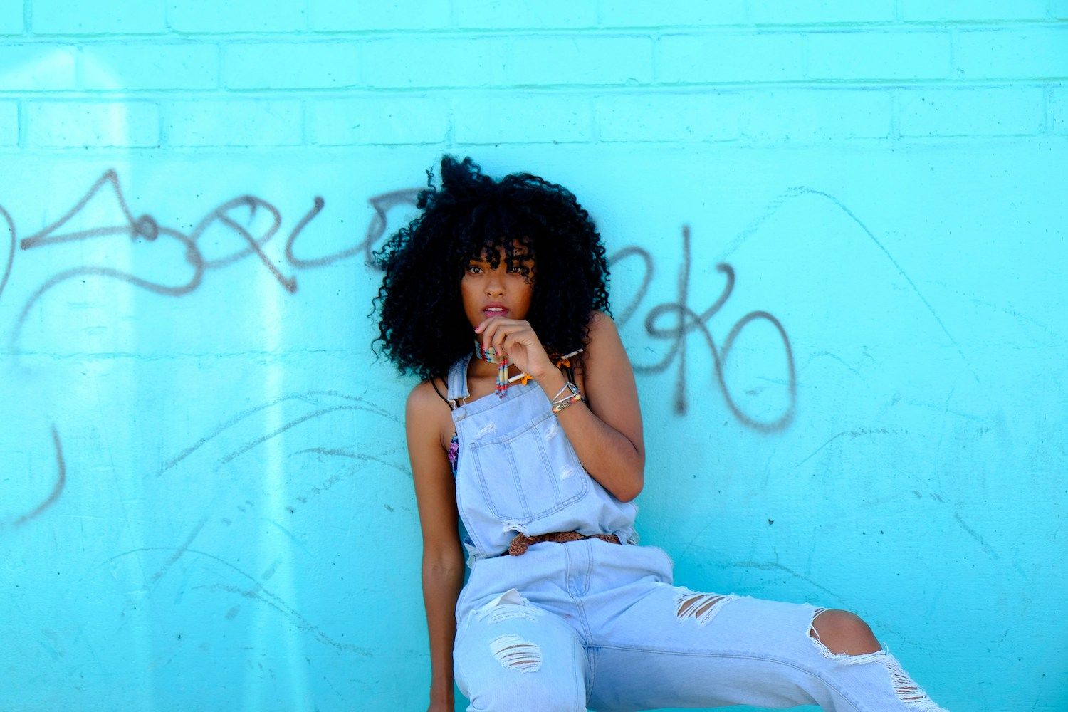Woman in denim overalls sits against a graffiti-covered brick wall