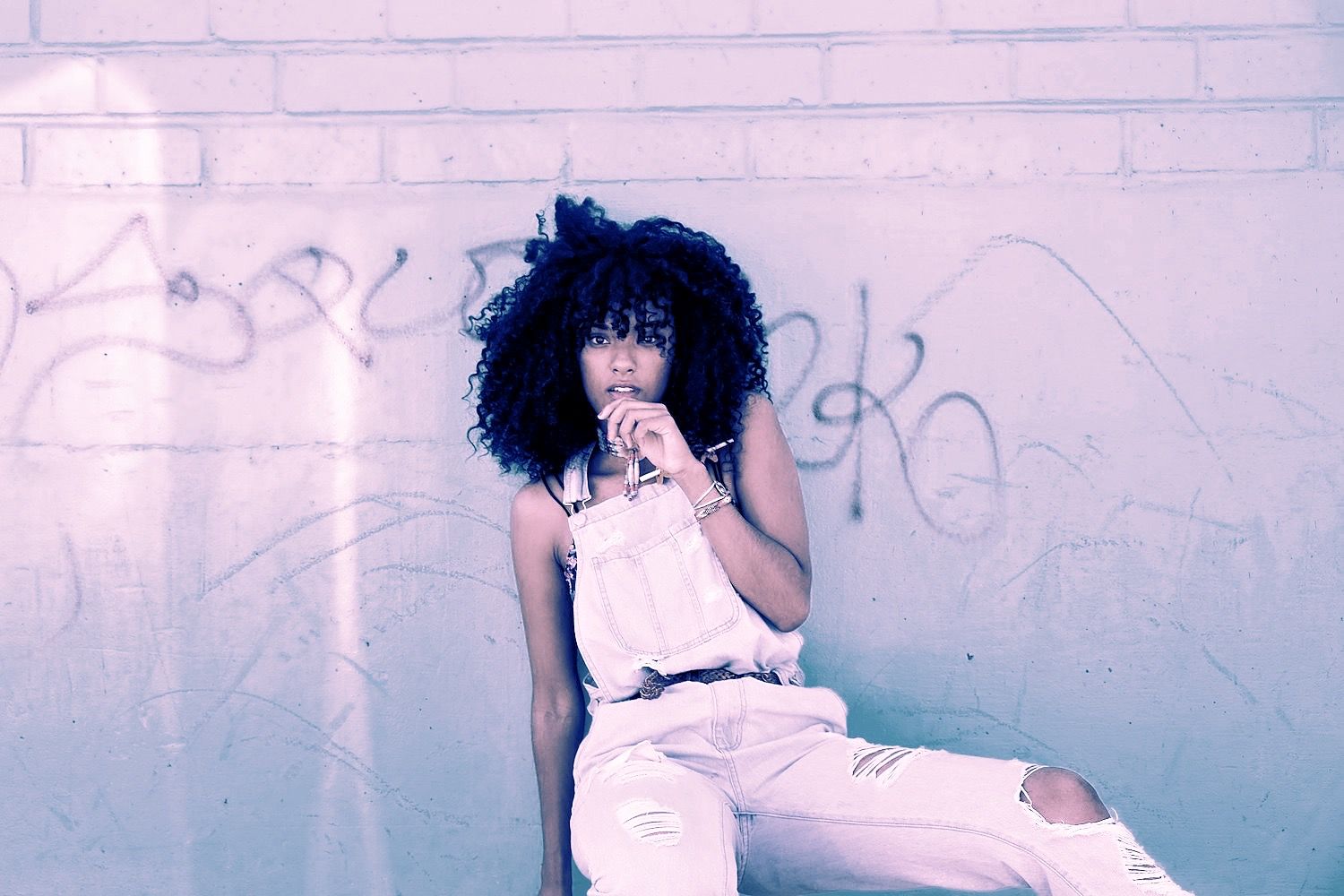Woman in overalls sits against a graffiti-covered brick wall