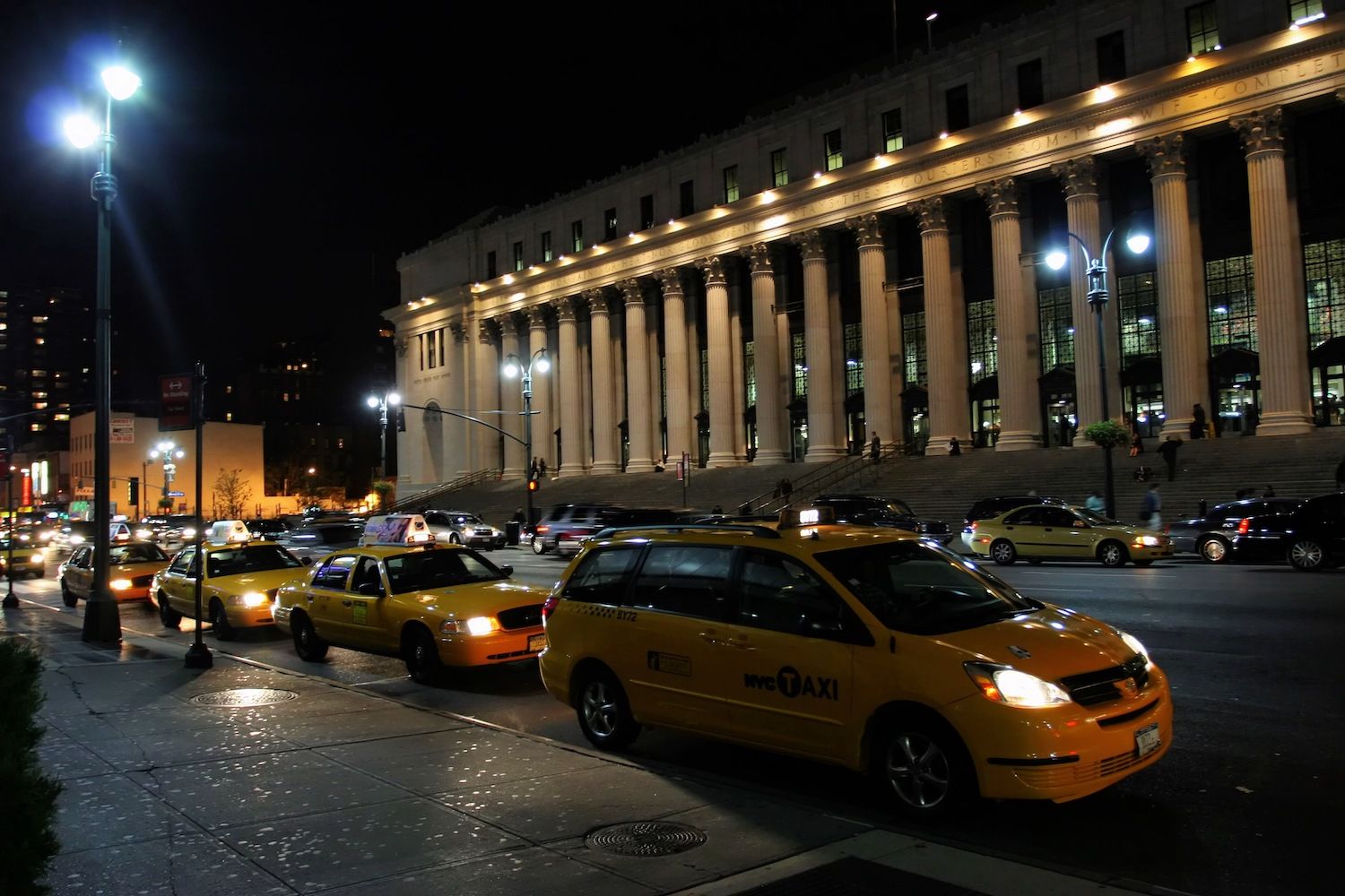 Yellow taxis line a busy street outside a columned building at night