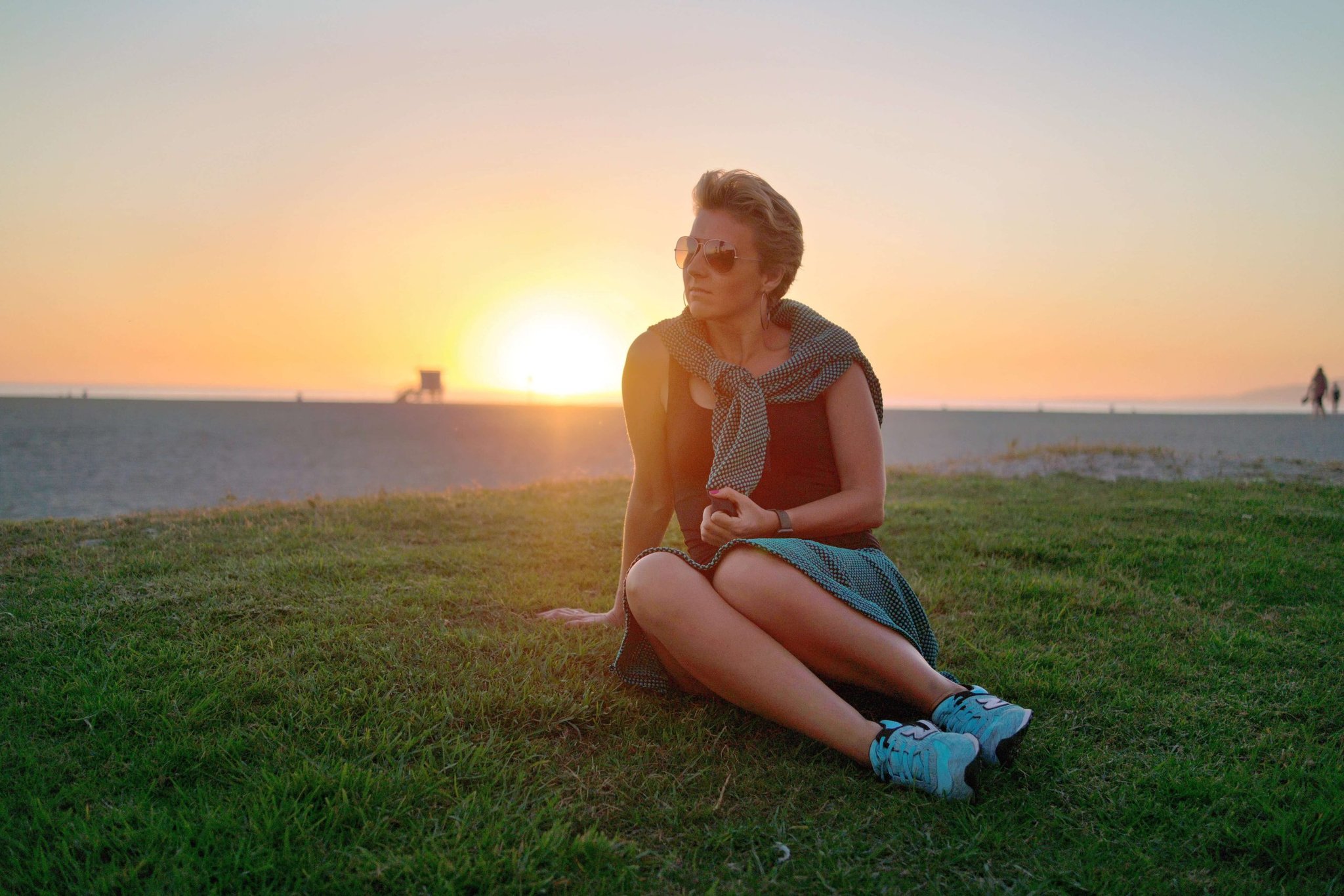 Woman wearing sunglasses sits on grass near a beach at sunset