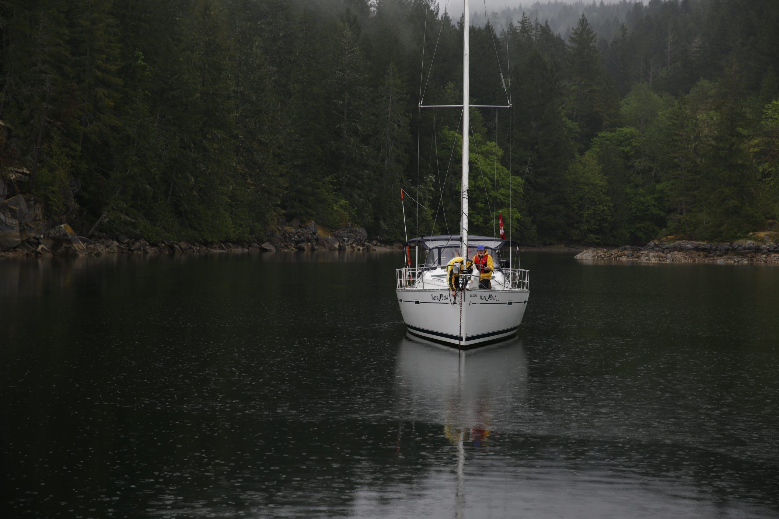 Two people stand aboard a sailboat in a forested inlet